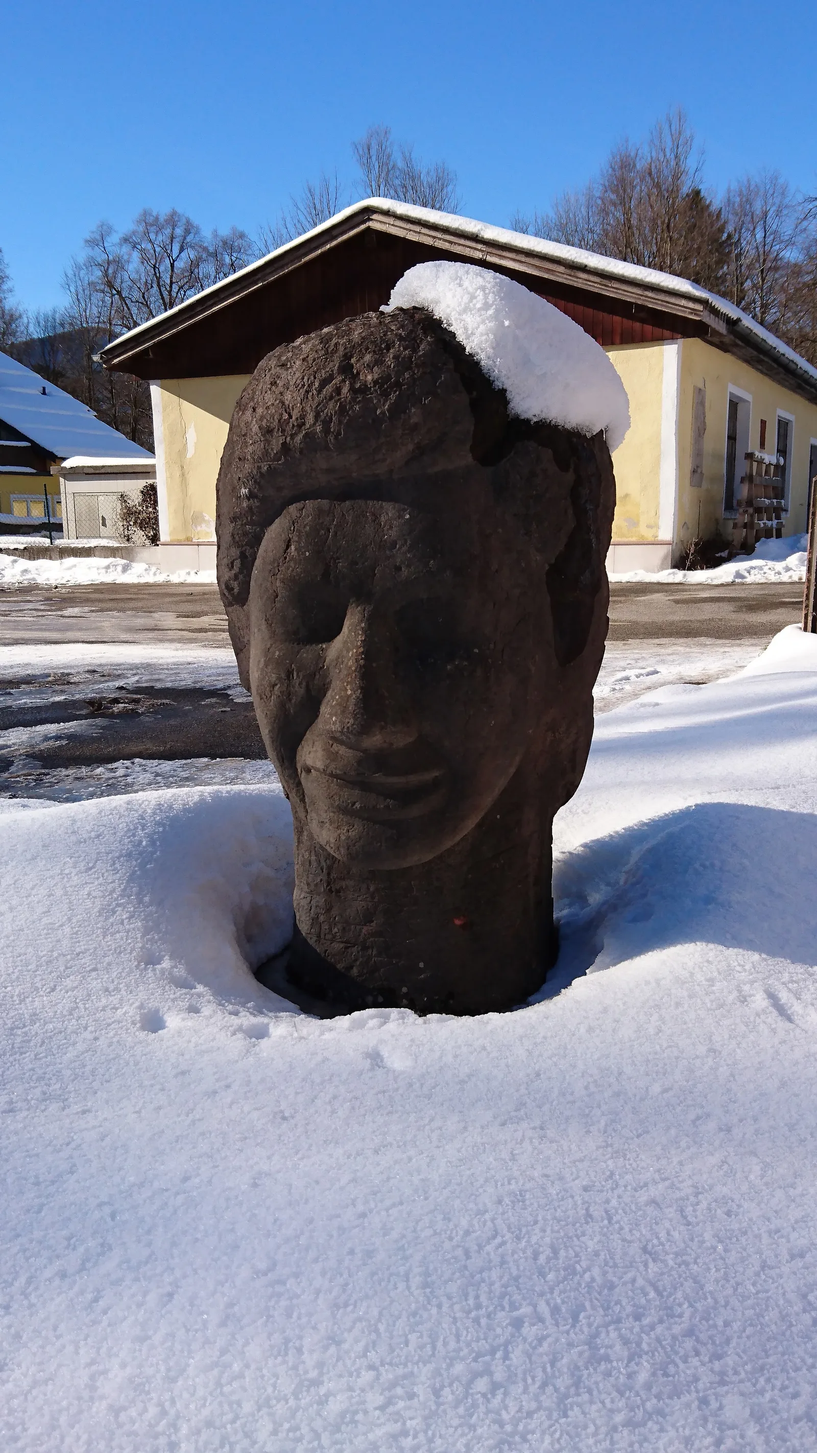Kopf aus rotbraunem Stein, von Hand gehauen, im Schnee vor dem Werksgebäude in Oberalm