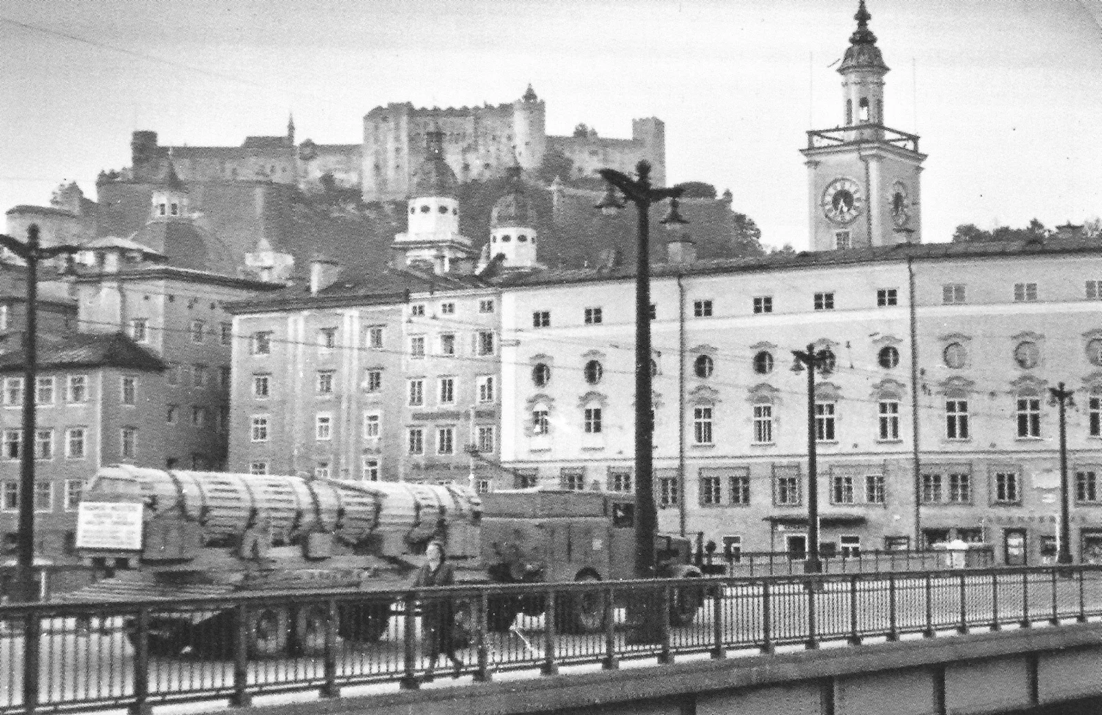 Historische Schwarzweißaufnahme eines Tiefladers mit einer langen Steinsäule auf der Salzburger Staatsbrücke, im Hintergrund die Festung